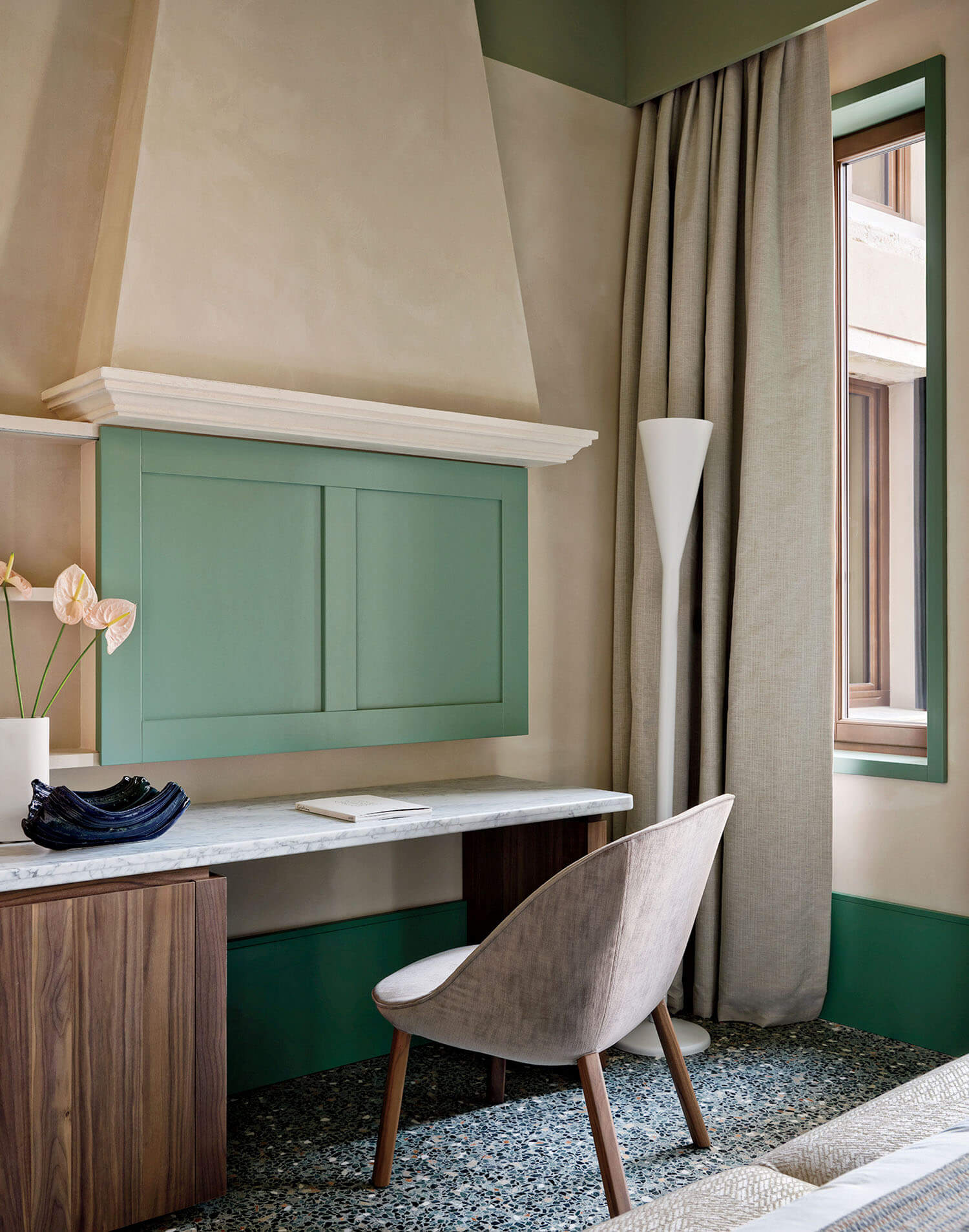 Modern workspace with a wooden desk, angled chair, green wall panel, floor lamp, vase with flowers, and large window with beige curtains.