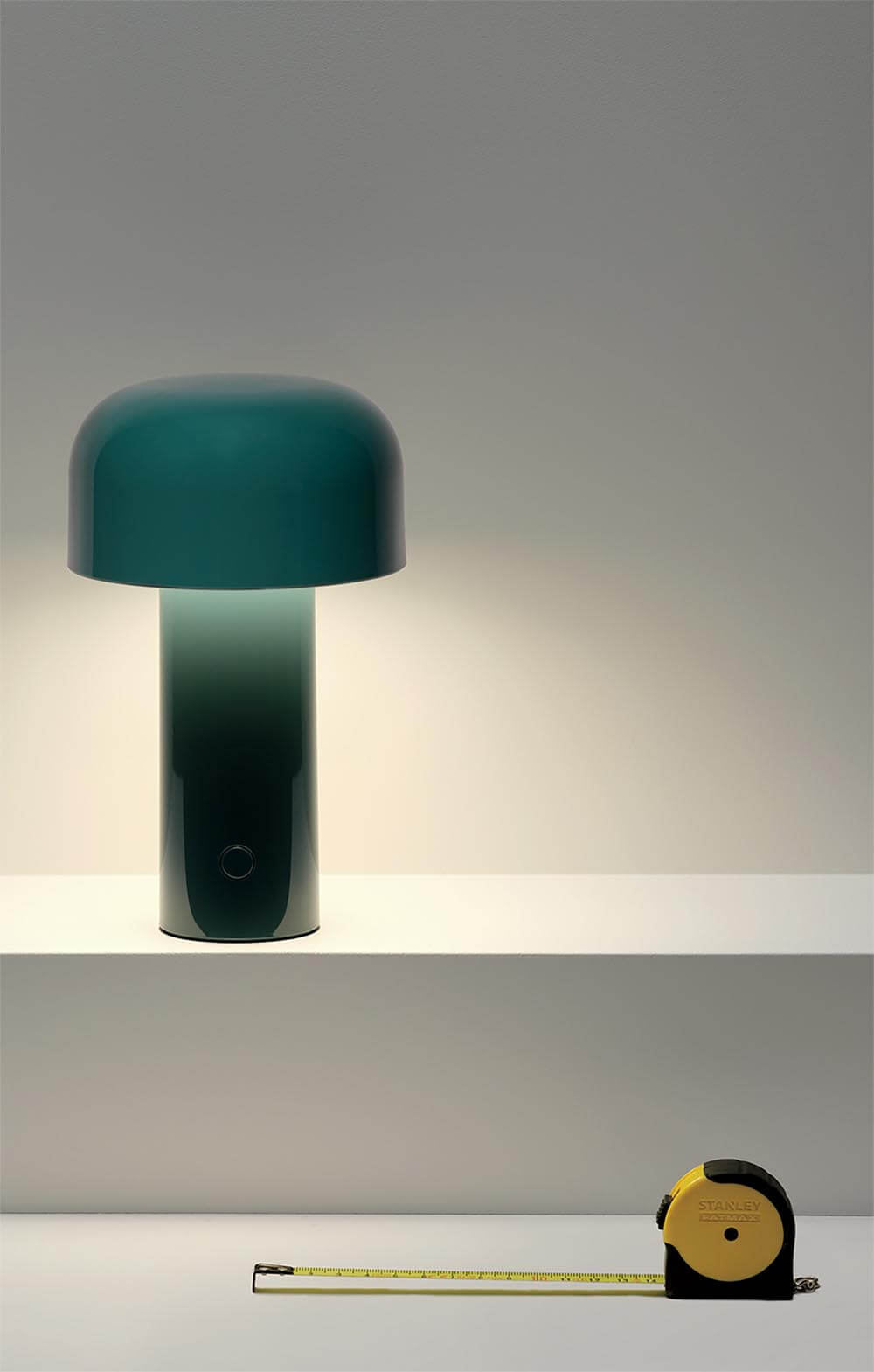 A green table lamp sits on a white shelf above a yellow and black tape measure extended across a lower surface.