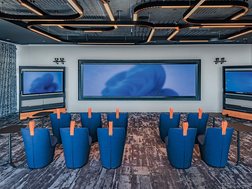 A modern conference room with blue chairs facing three large display screens on the front wall, small tables beside the chairs, and unique ceiling lighting.
