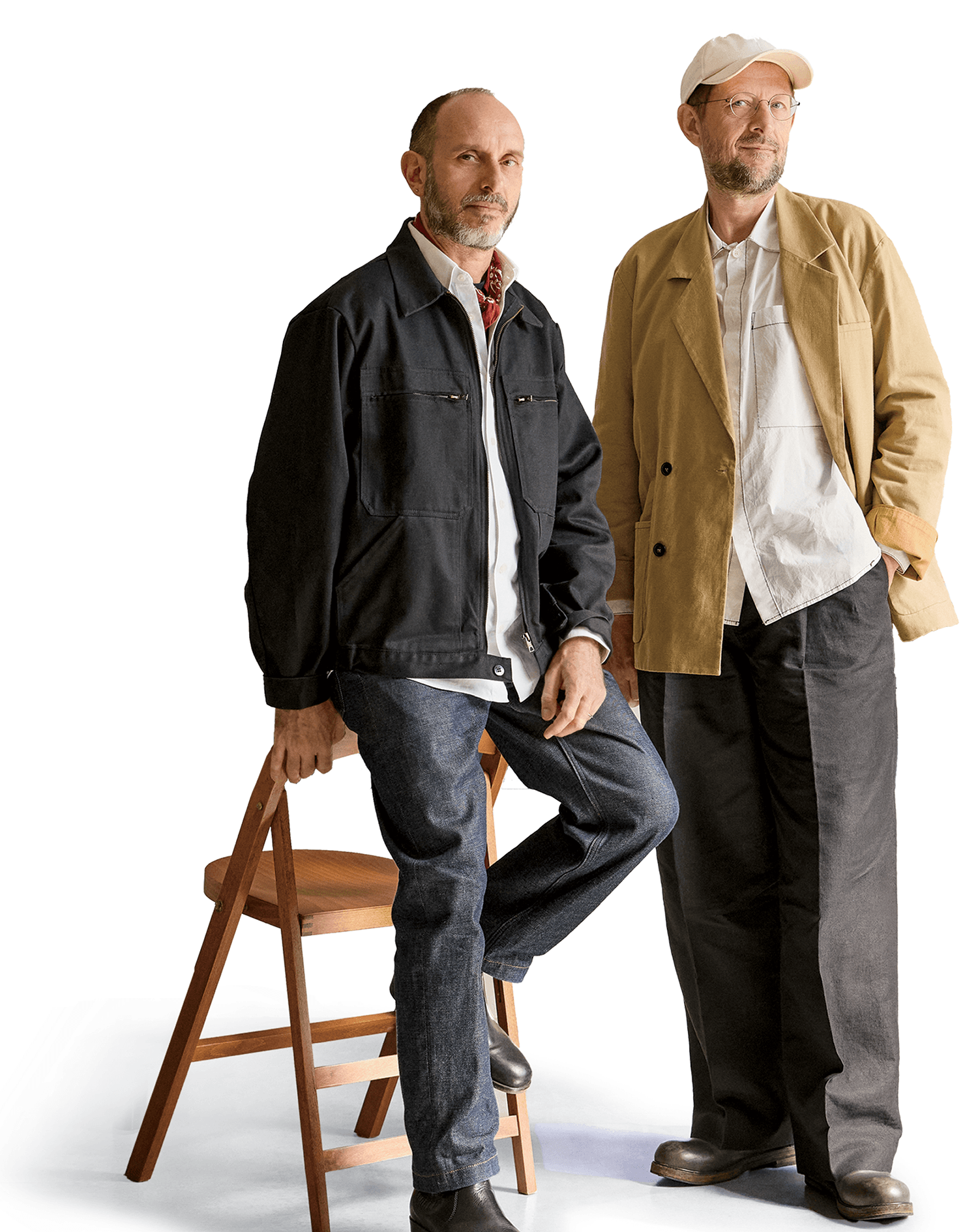 Two men pose against a white background; one is sitting on a wooden chair, wearing a black jacket and jeans, while the other stands, wearing a beige jacket and dark pants.