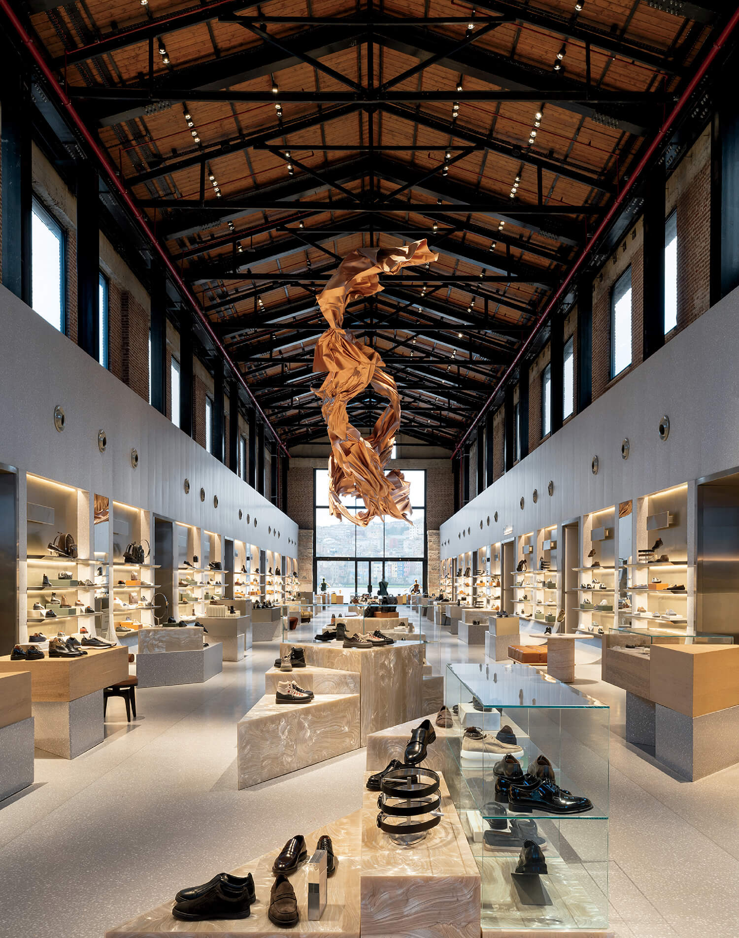 Spacious shoe store with high vaulted ceiling, skylights, and a central abstract wooden sculpture. Shoes are displayed on various platforms throughout the space.