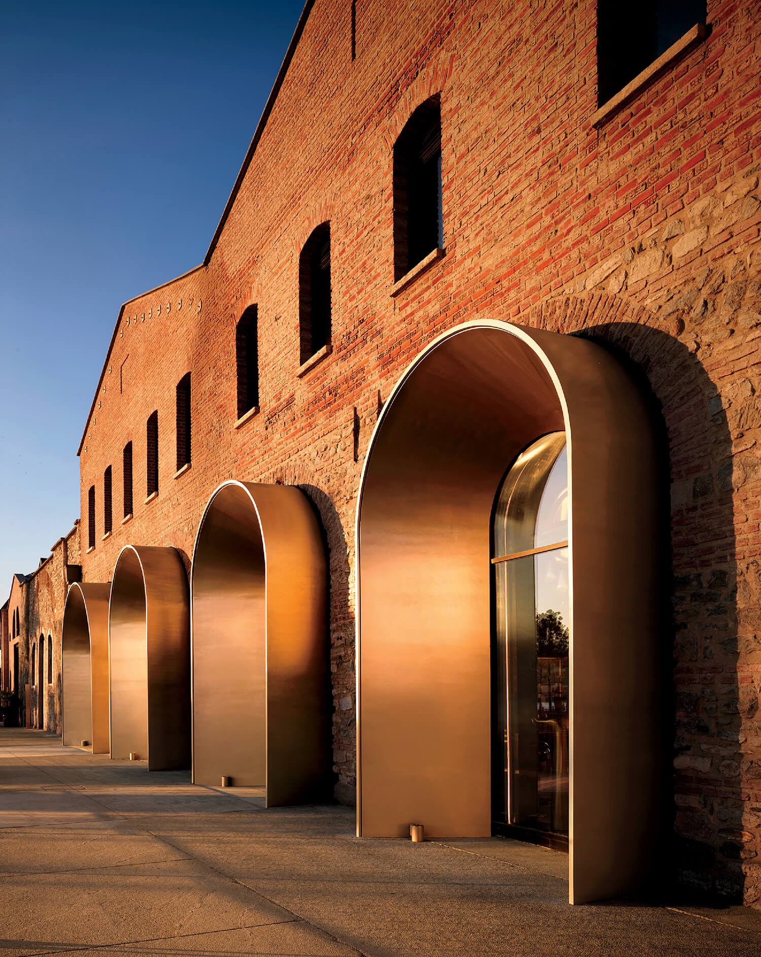 A brick building with tall, arched bronze doorways reflecting sunlight, featuring rectangular windows above each arch.