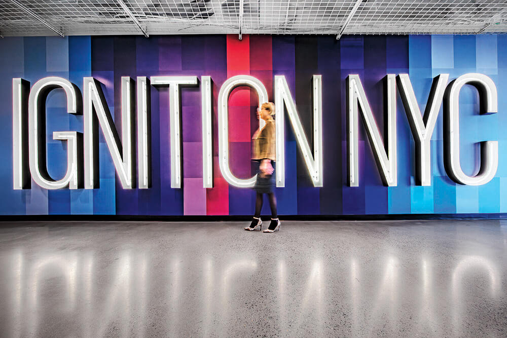 A person walks past a neon sign reading “IGNITION NYC” on a blue and purple striped wall in a modern indoor space.
