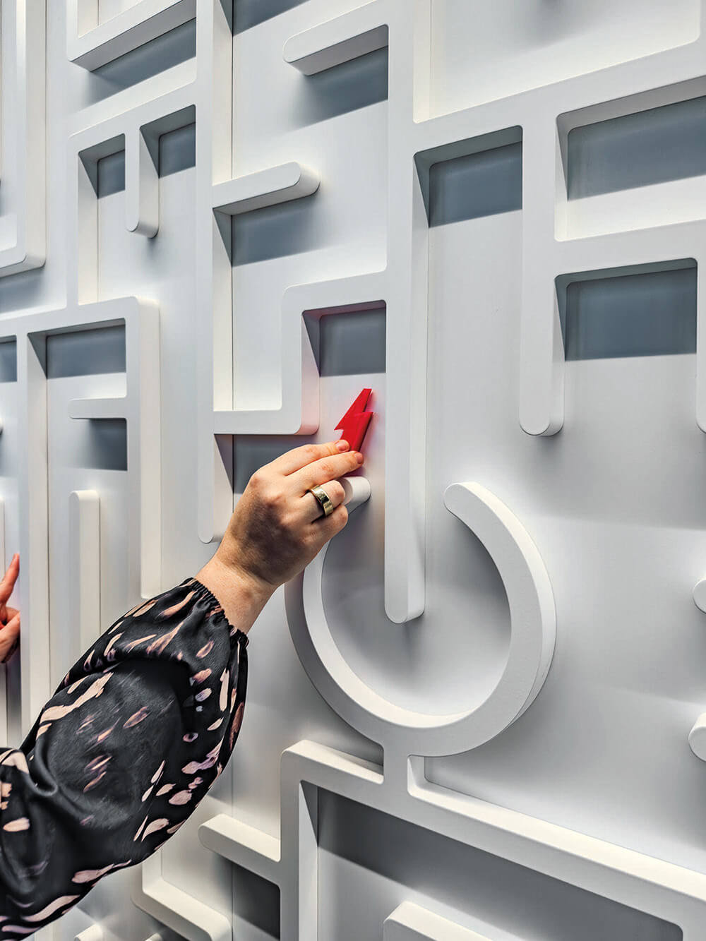 A person in a patterned sleeve slides a small red object along a white, geometric maze-like wall feature.