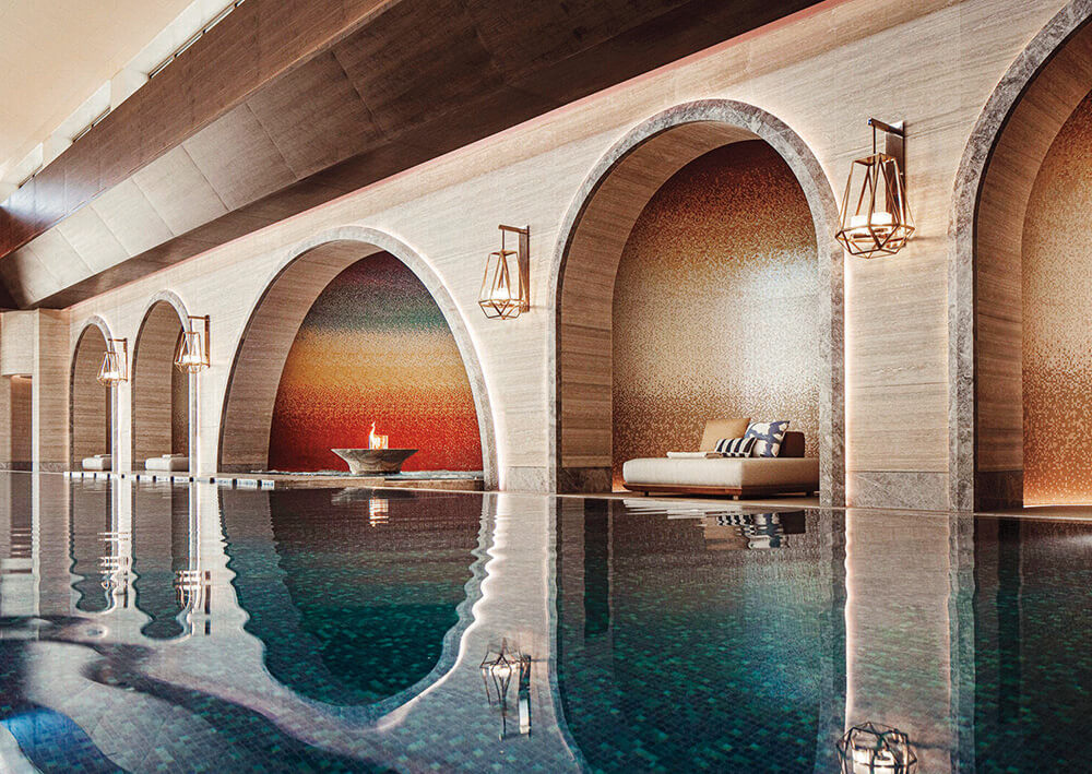 Indoor pool area with arched alcoves, lounge chairs, mosaic tile walls, lantern-style wall lights, and a decorative fire bowl beside the pool.