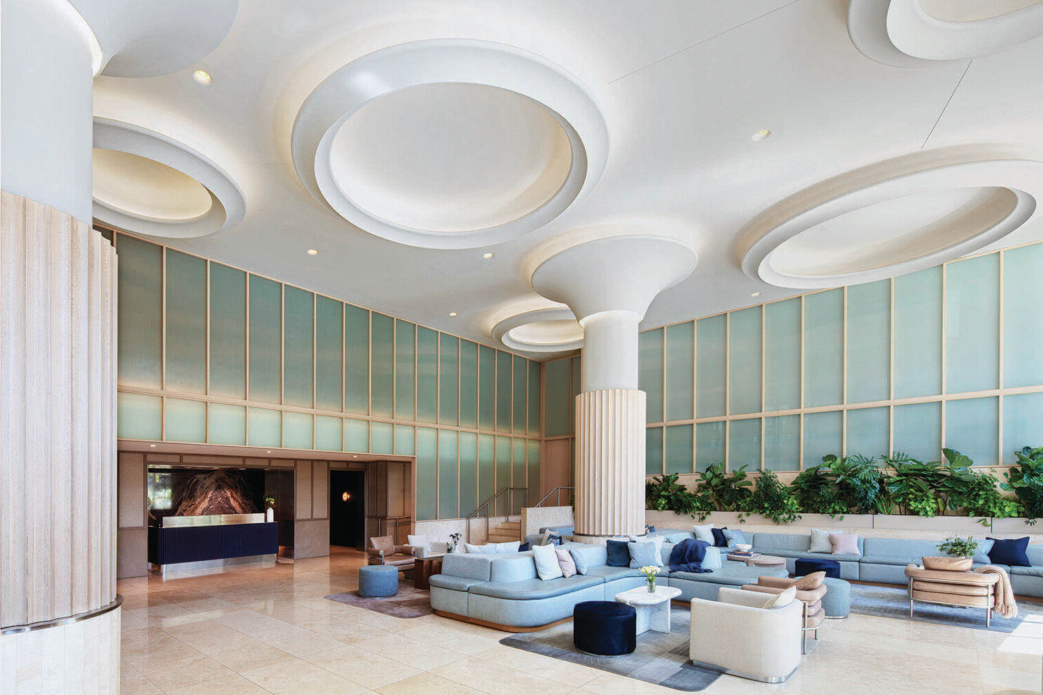 Modern hotel lobby with high ceilings, circular ceiling features, light-colored furniture, indoor plants, and a reception desk on the left.