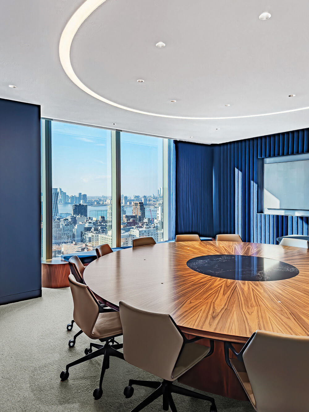 Modern conference room with a large oval wooden table, office chairs, blue accent walls, and floor-to-ceiling windows overlooking a city skyline.