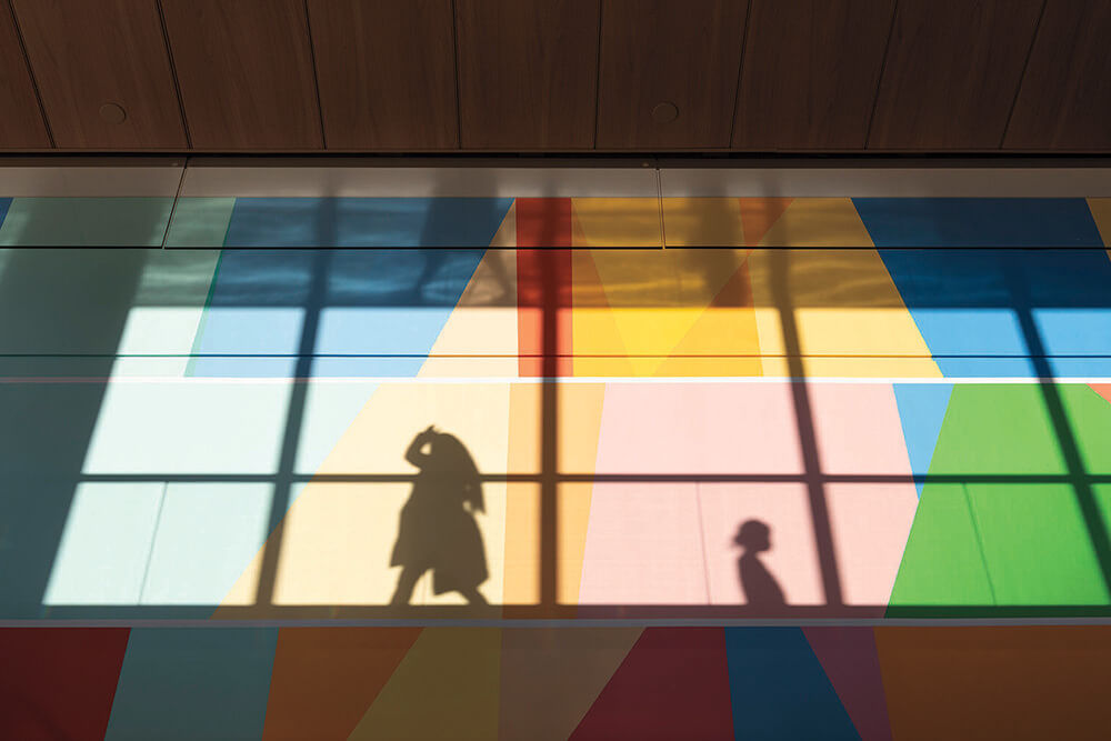 Shadows of two people cast on a colorful geometric wall with sunlight streaming through large windows above.