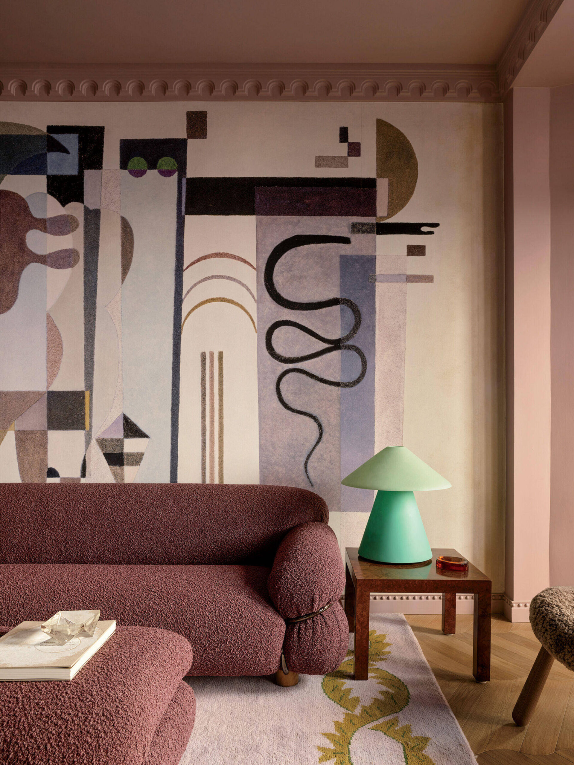 A living room with a textured maroon sofa, a green lamp on a wooden side table, and a large abstract mural on the wall.