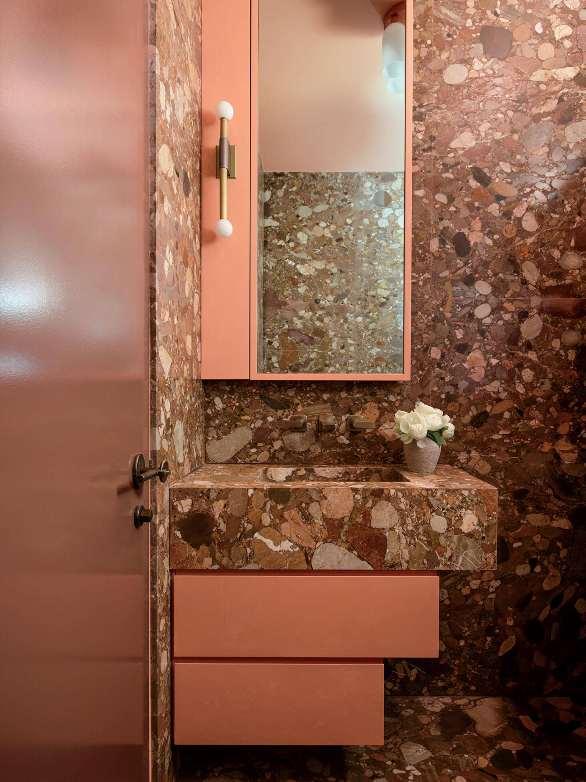 Small bathroom with pink terrazzo walls and sink, pink floating drawers, a mirrored cabinet, and a small white vase with flowers on the counter.