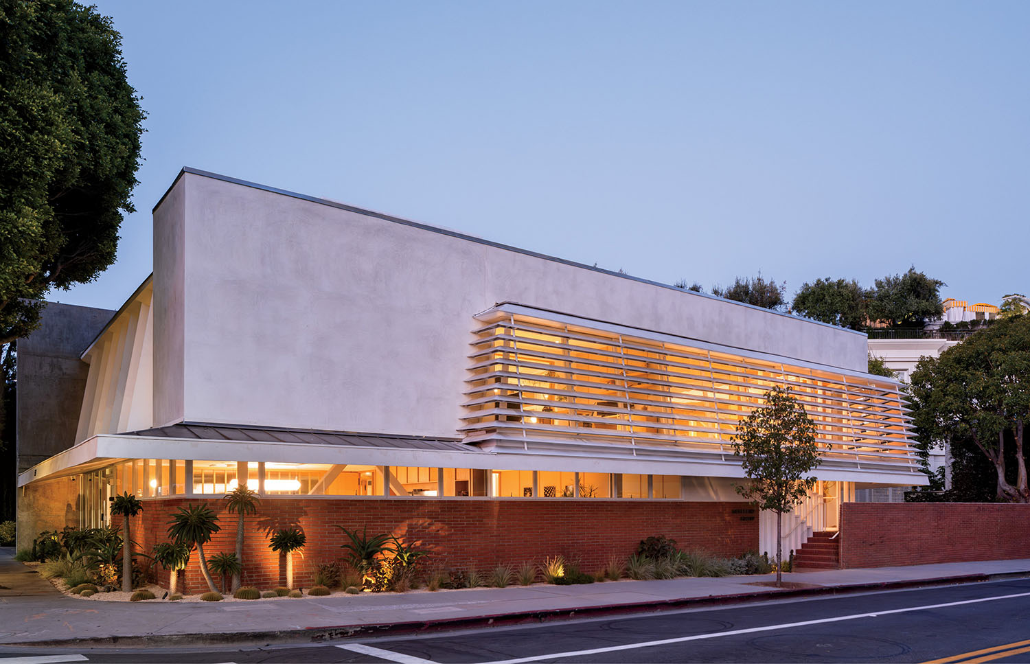 Modern two-story building with white walls, horizontal slats, large windows, and brick base, surrounded by plants and trees, photographed at dusk.