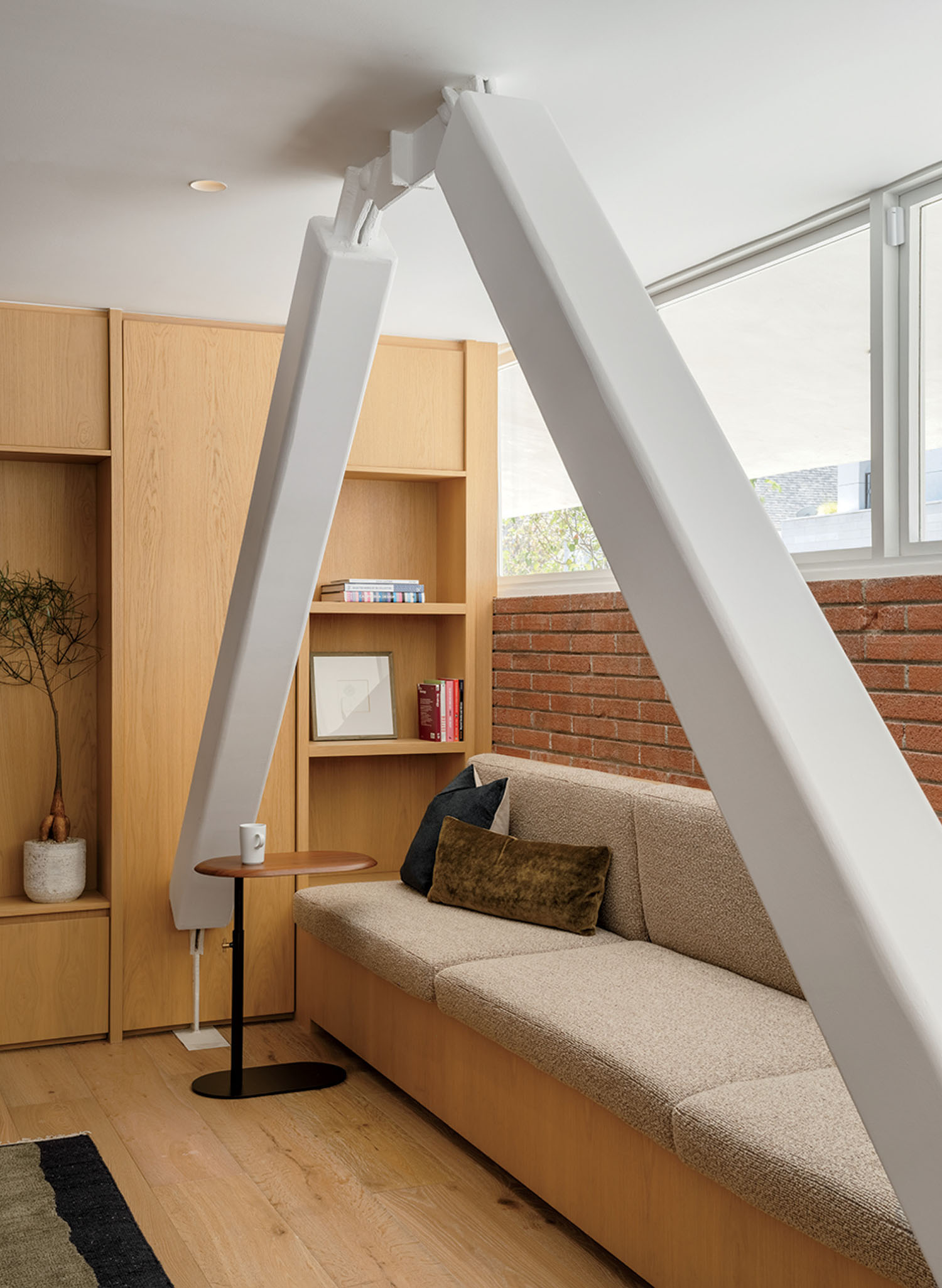 Modern living room with a beige sofa, wooden shelves, a small round side table, and a large white structural beam angled over the seating area.