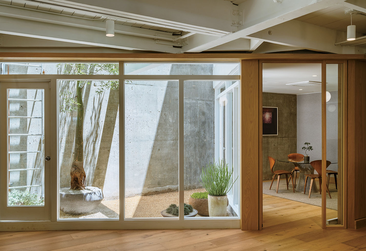 Modern interior with large glass windows showing a small courtyard garden; adjacent room has a table, chairs, and wall art. Natural light fills the space.