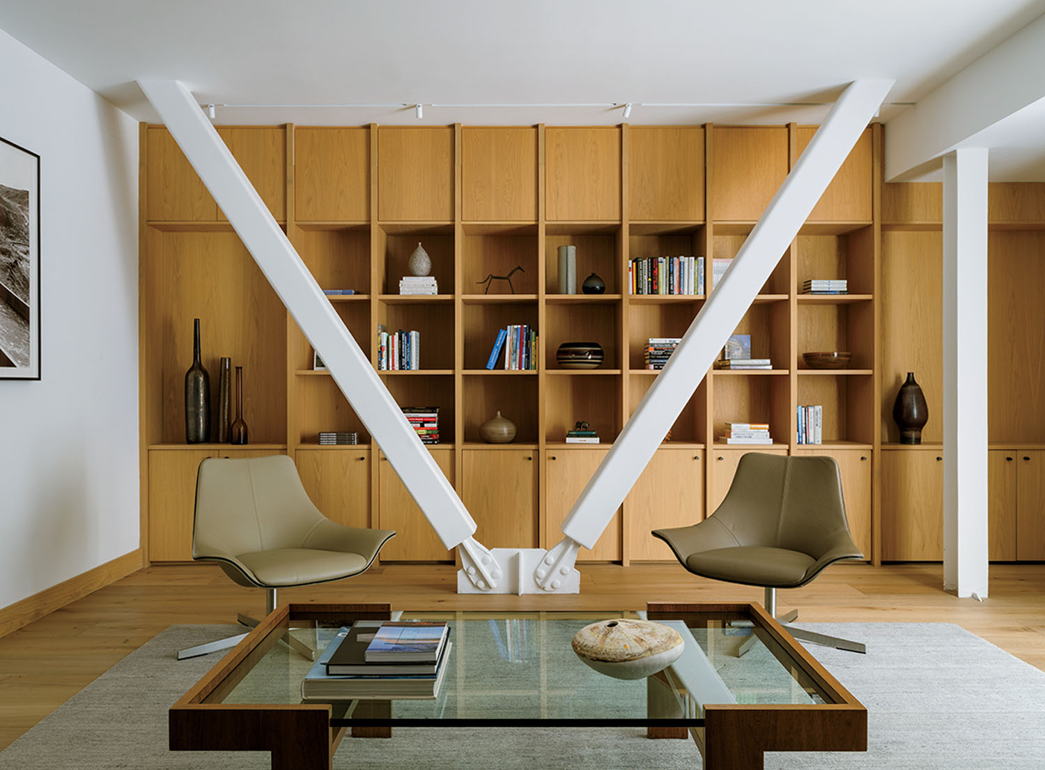 Modern living area with two armchairs, a glass coffee table, and a wall of wooden shelves, divided by two large white diagonal support beams.