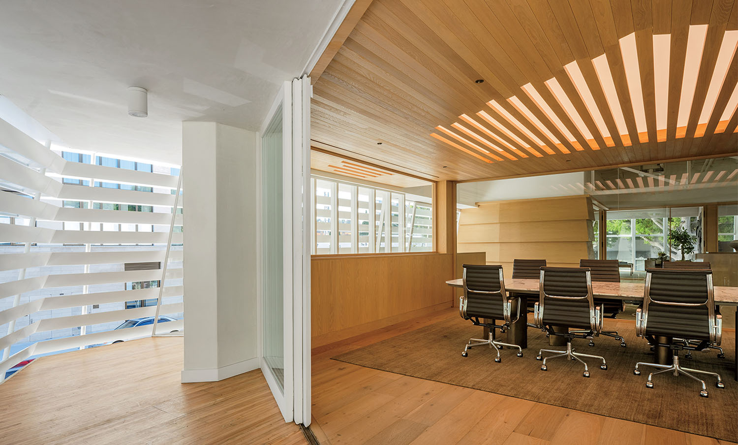 Modern office space with a glass-walled conference room, wooden floors and ceiling, and ergonomic chairs around a long table. Natural light enters through large windows with horizontal blinds.