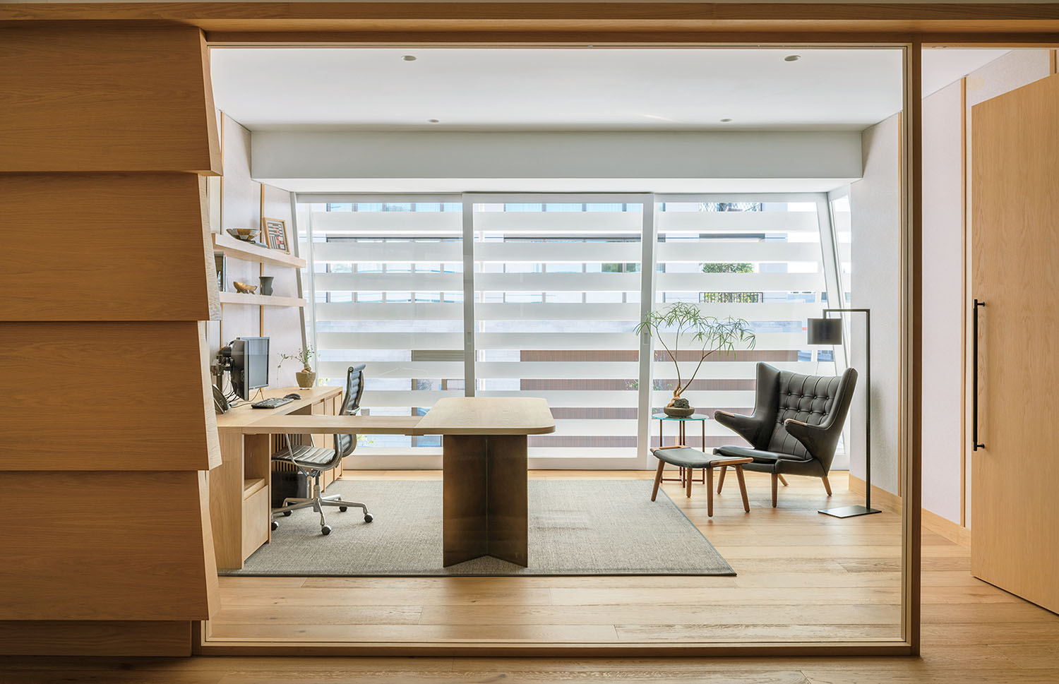 Modern office with a wooden desk, office chair, armchair, floor lamp, small table, shelves, and large windows with horizontal blinds.