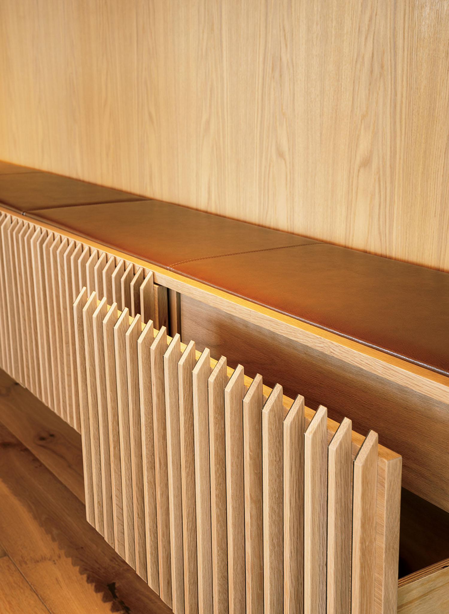Wooden cabinet with vertical slatted doors and cushioned brown leather top, set against a matching wood wall and floor.