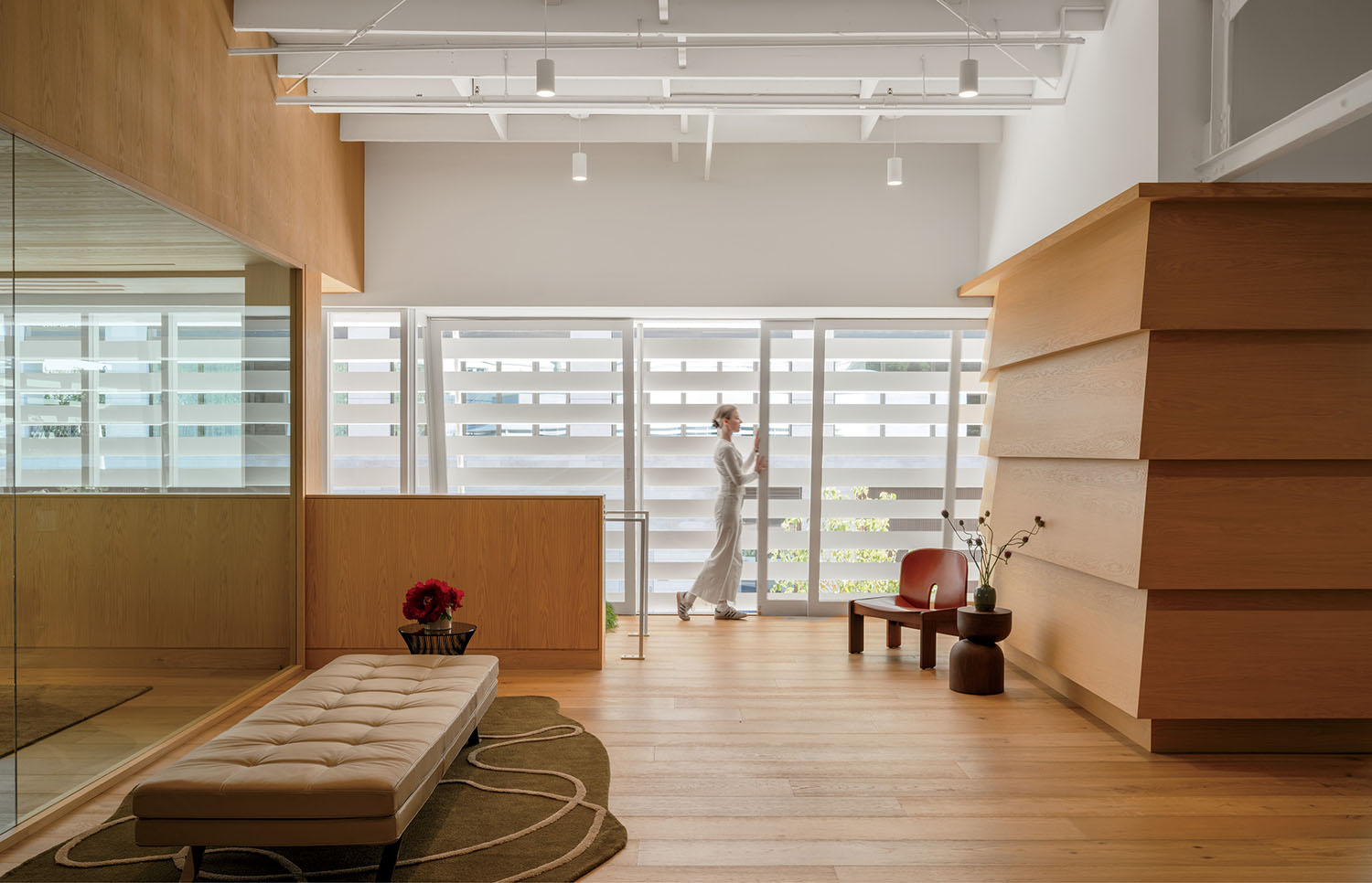 A person walks past large windows in a modern, minimalist interior with wooden floors, furniture, and a neutral color palette.
