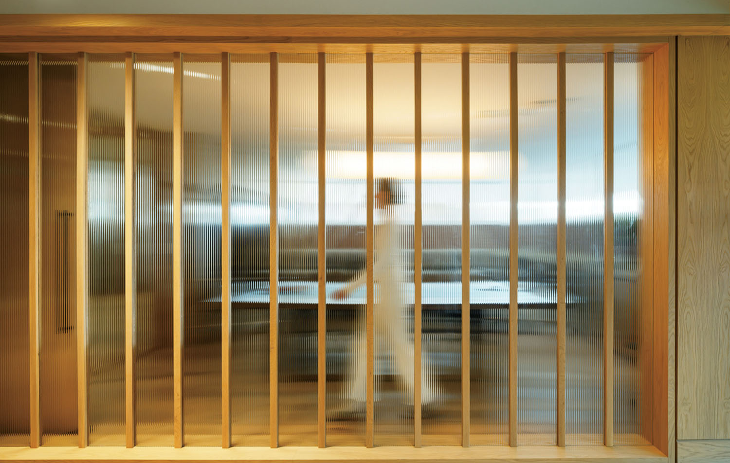 A person in white walks behind a translucent glass wall with vertical wooden slats in a modern interior space.