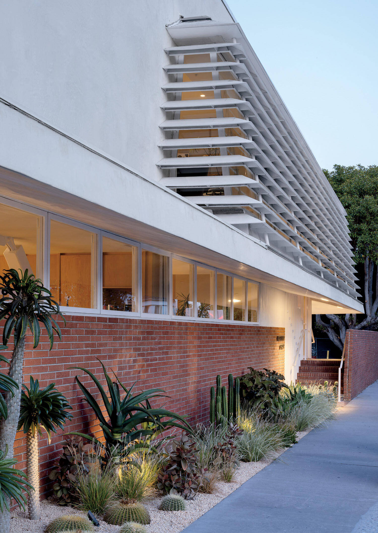 Modern building with a brick lower facade, white upper level with horizontal louvered sunshades, and desert landscaping featuring cacti and succulents along the sidewalk.