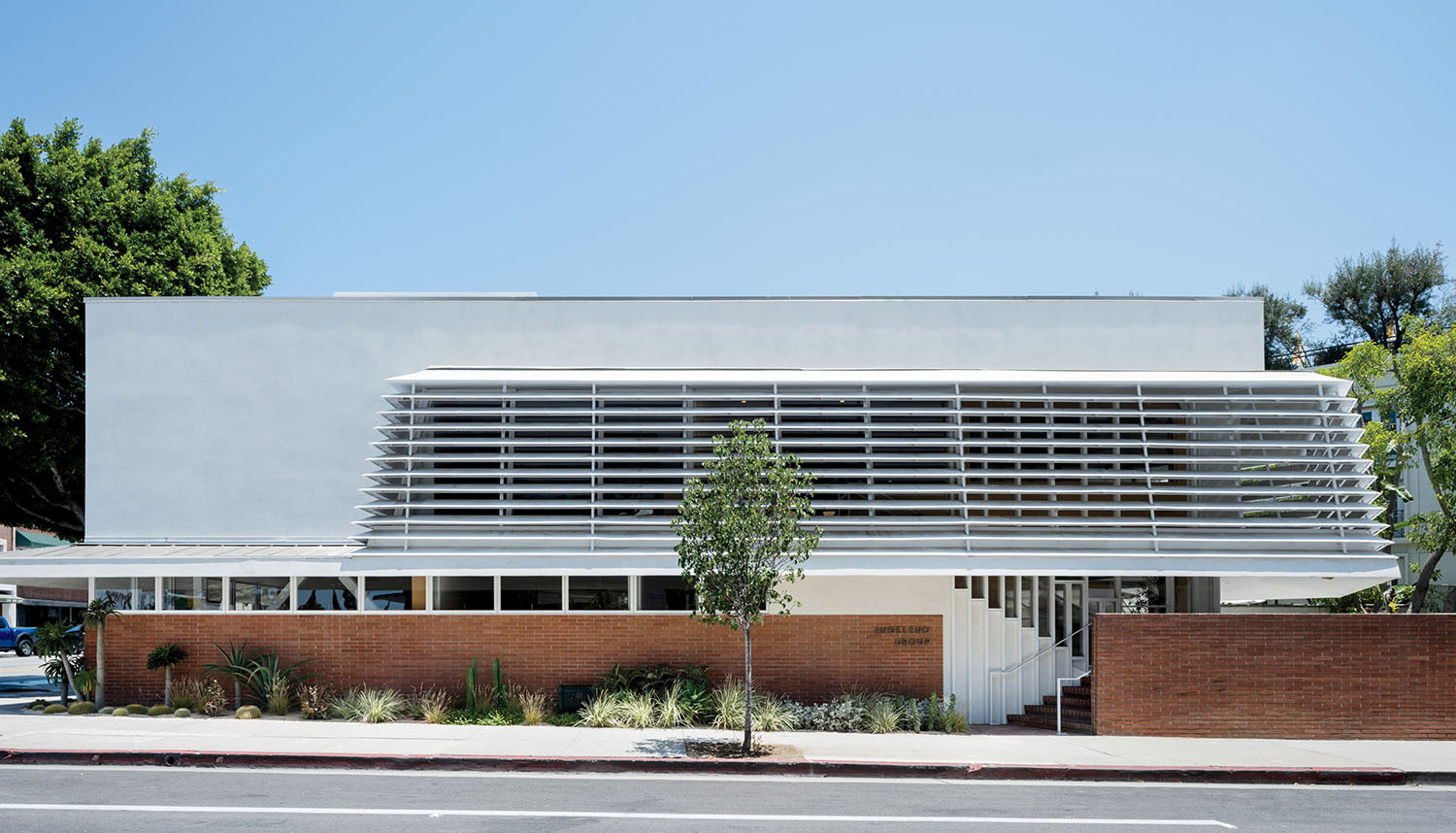 A modern building with white horizontal louvers, a brick base, and landscaping in front under a clear blue sky.