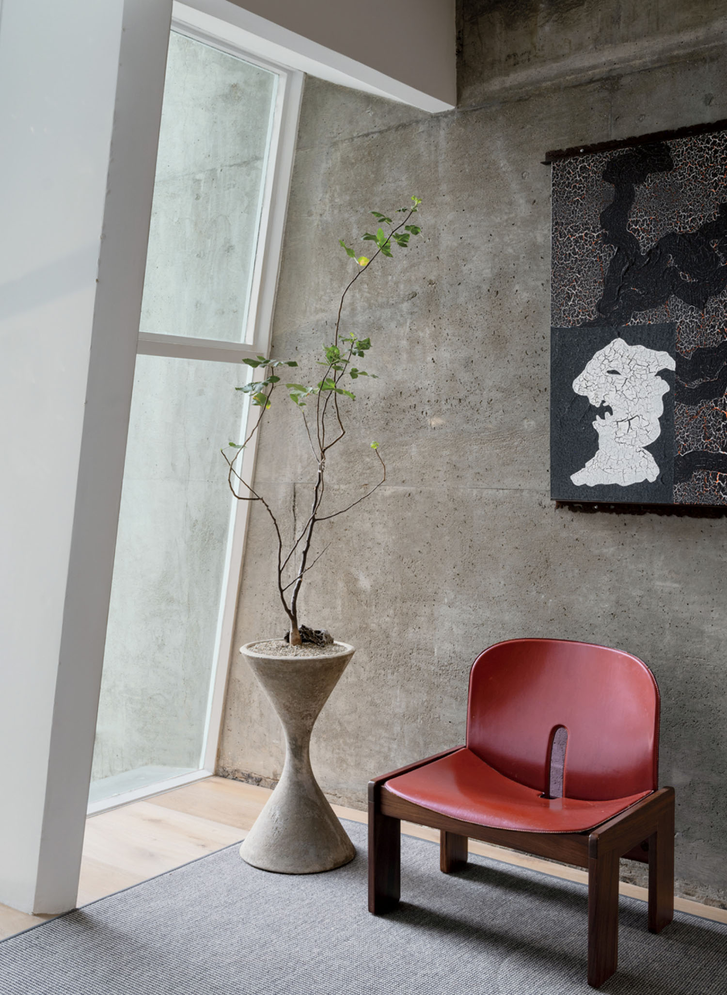 A modern room with a red chair, a tall potted plant, a gray rug, and abstract artwork on a concrete wall near a floor-to-ceiling window.