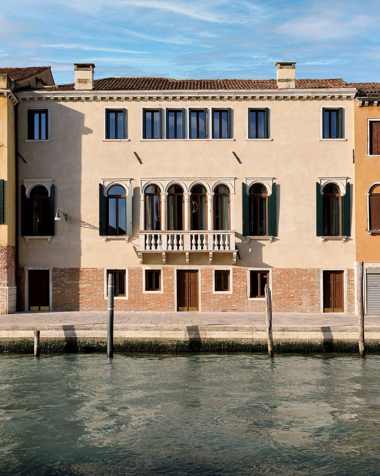 A beige, three-story building with arched windows and balcony faces a canal with wooden poles in front under a blue sky.