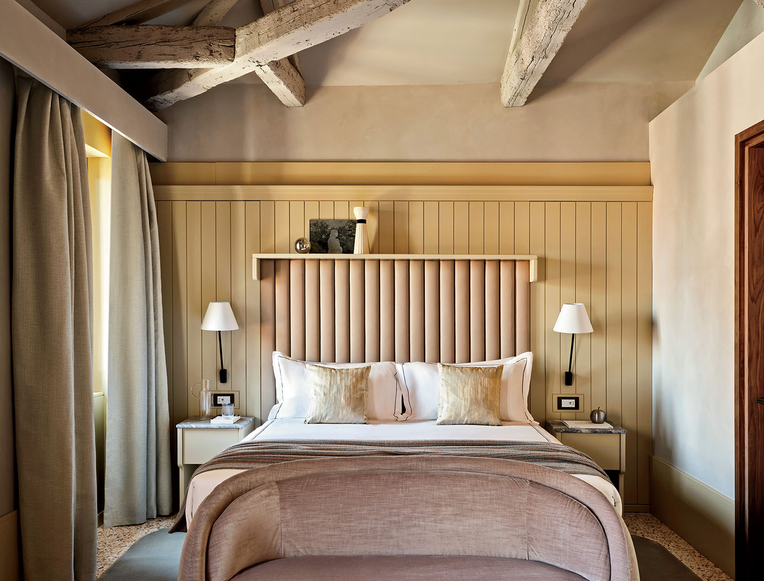 Neutral-toned bedroom with a double bed, beige upholstered headboard, two bedside tables with lamps, exposed wooden beams, and large curtains covering a window.