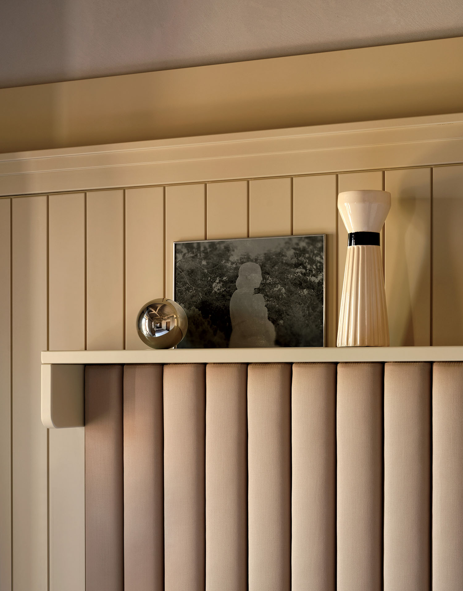 A shelf displays a round metallic object, a framed black-and-white photo, and a tall cream-colored vase against beige vertical wall panels.