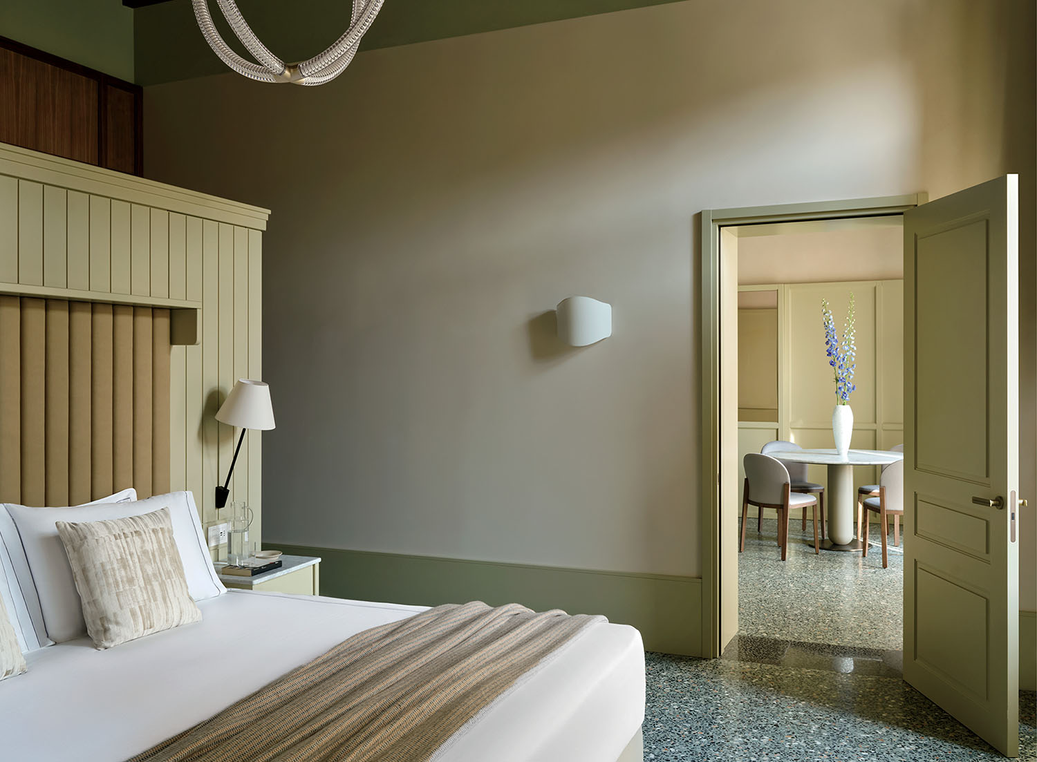 A modern bedroom with a large bed, neutral tones, and an open door showing a dining area with a round table, four chairs, and a tall vase with flowers.