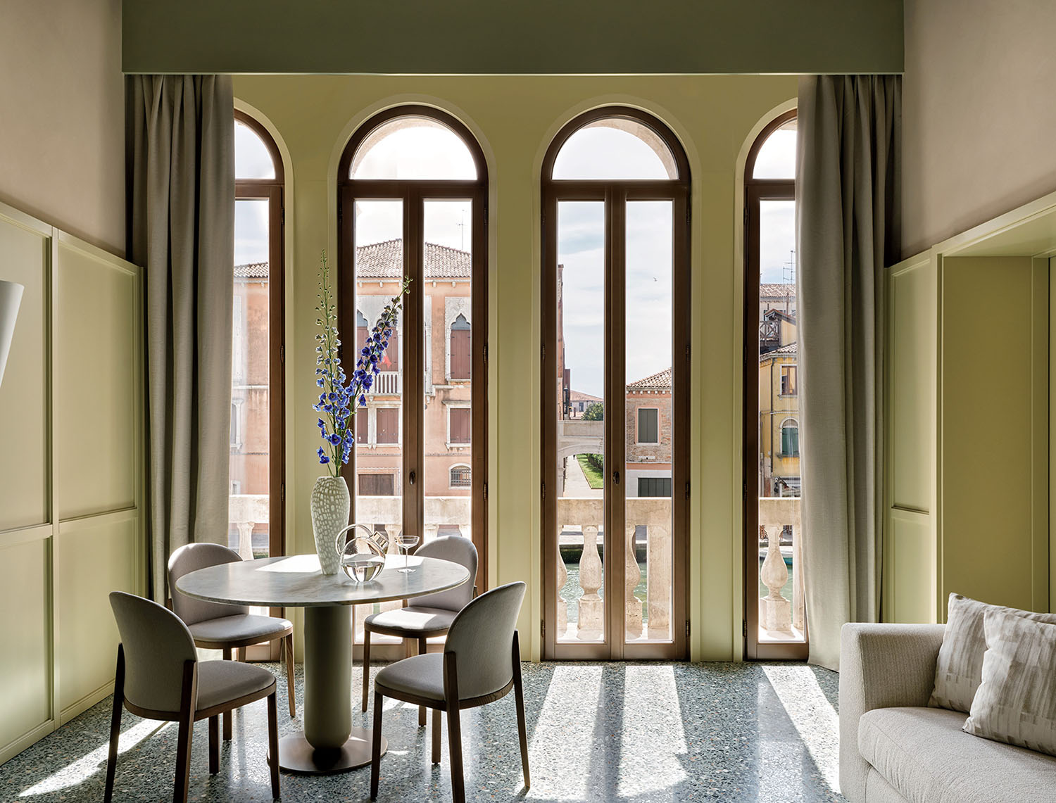 Modern dining area with round table and four chairs near tall arched windows, offering a view of historic buildings and a canal outside. Curtains frame the windows, and a vase decorates the table.