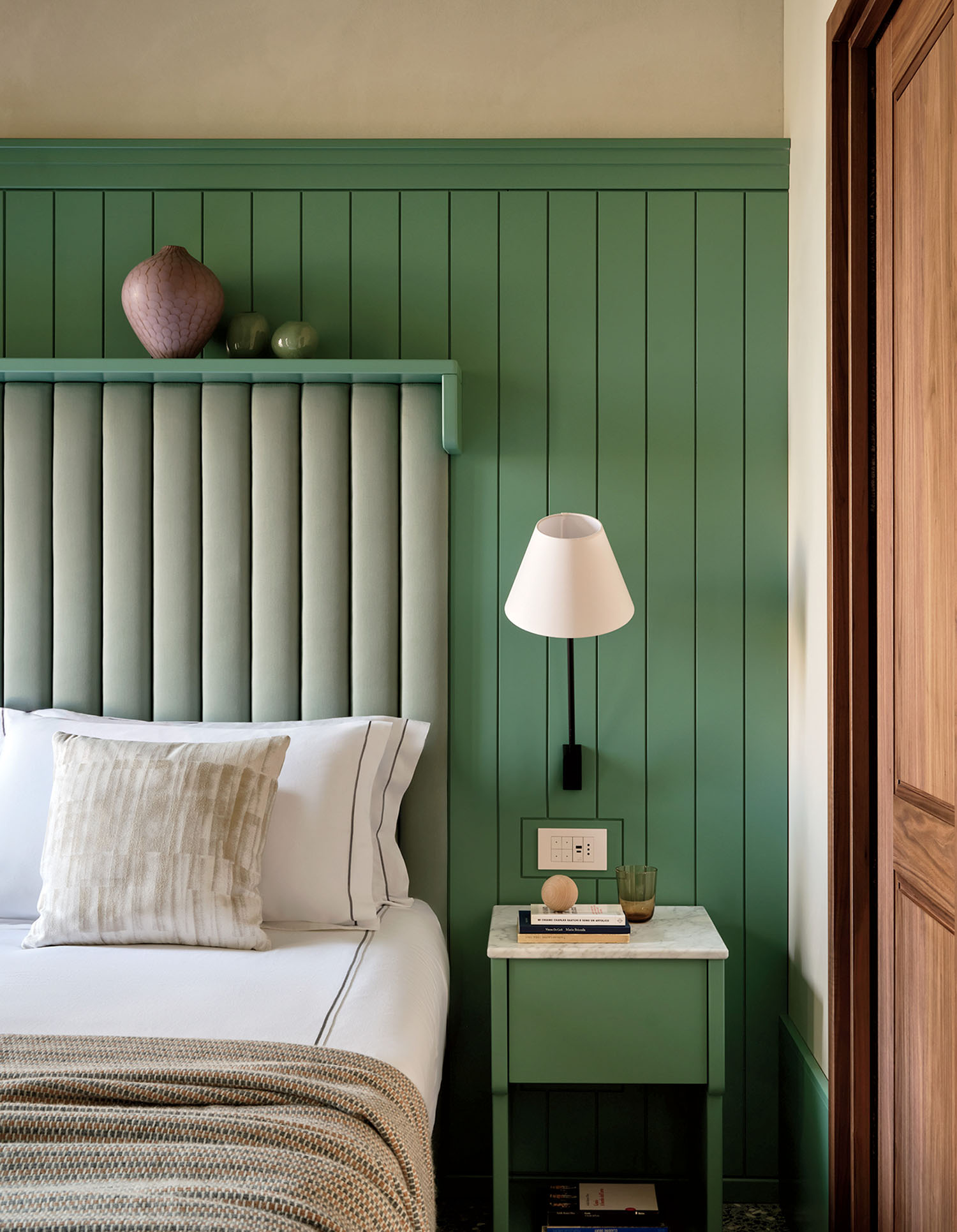A neatly made bed with white linens beside a green nightstand, a wall lamp, and a paneled green accent wall with decorative vases on a shelf.