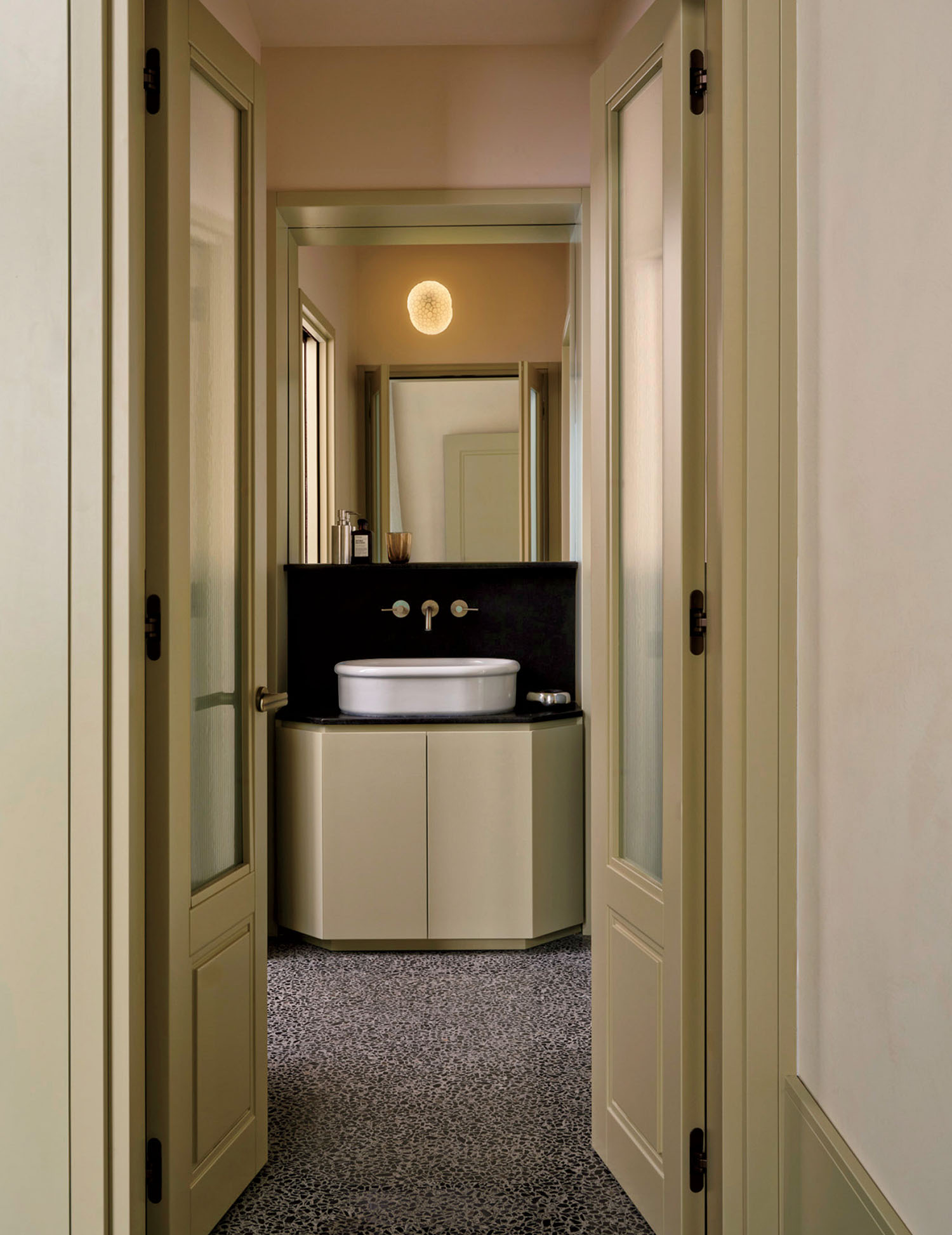 Narrow hallway with frosted glass doors leads to a small bathroom featuring a round sink, wall-mounted faucets, a mirror, and terrazzo flooring.