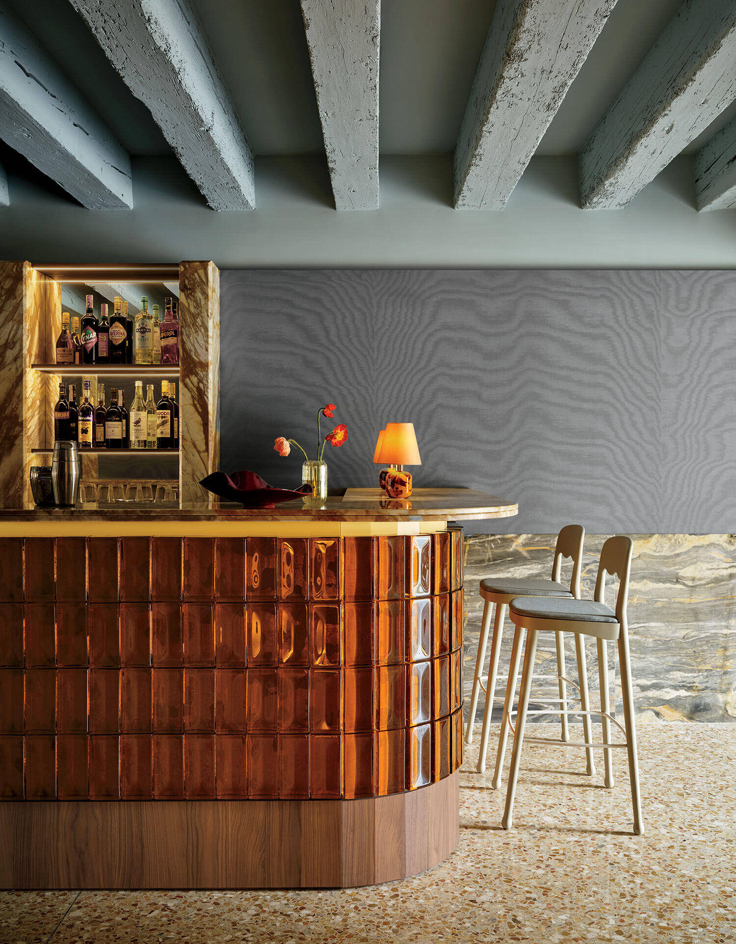 A modern bar with a copper-tiled counter, wooden bar stools, a shelf of liquor bottles, and a lamp on the counter, set against a textured gray wall and exposed ceiling beams.