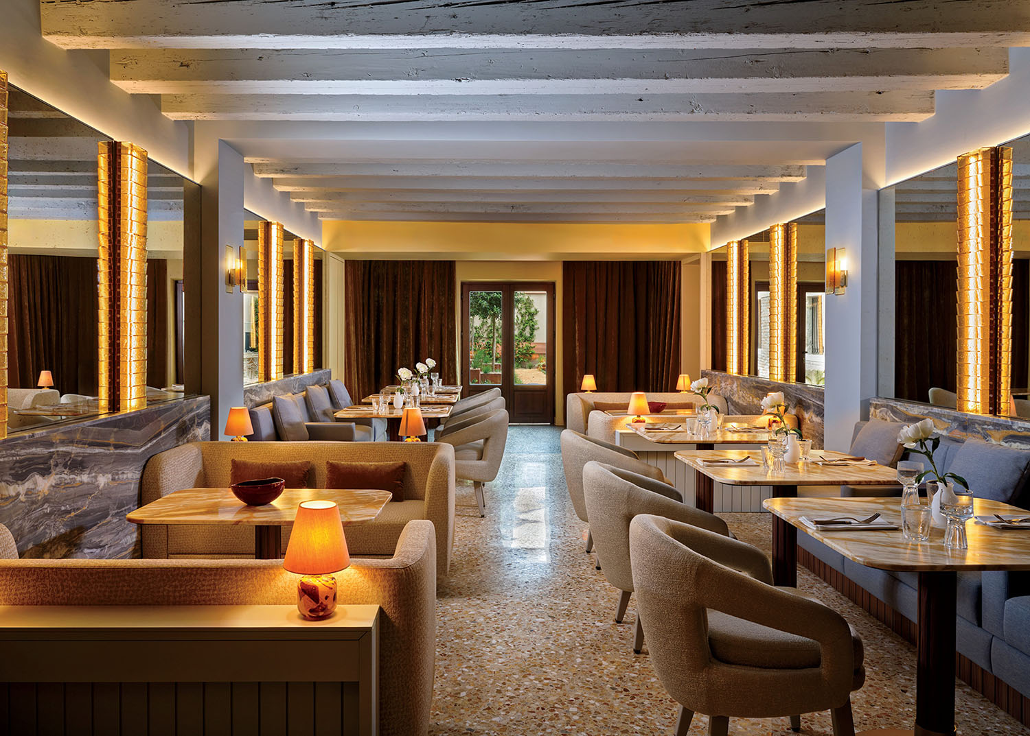 Modern restaurant interior with beige armchairs, wooden tables, soft lighting, decorative mirrors, and large windows at the far end.