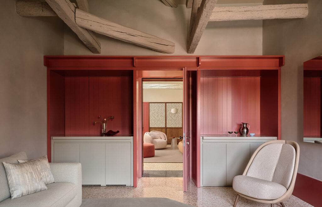 Modern interior with exposed wooden beams, red alcoves, a beige armchair, and a light sofa. Open doorway leads to another stylish room with soft pink tones and textured wall panels.