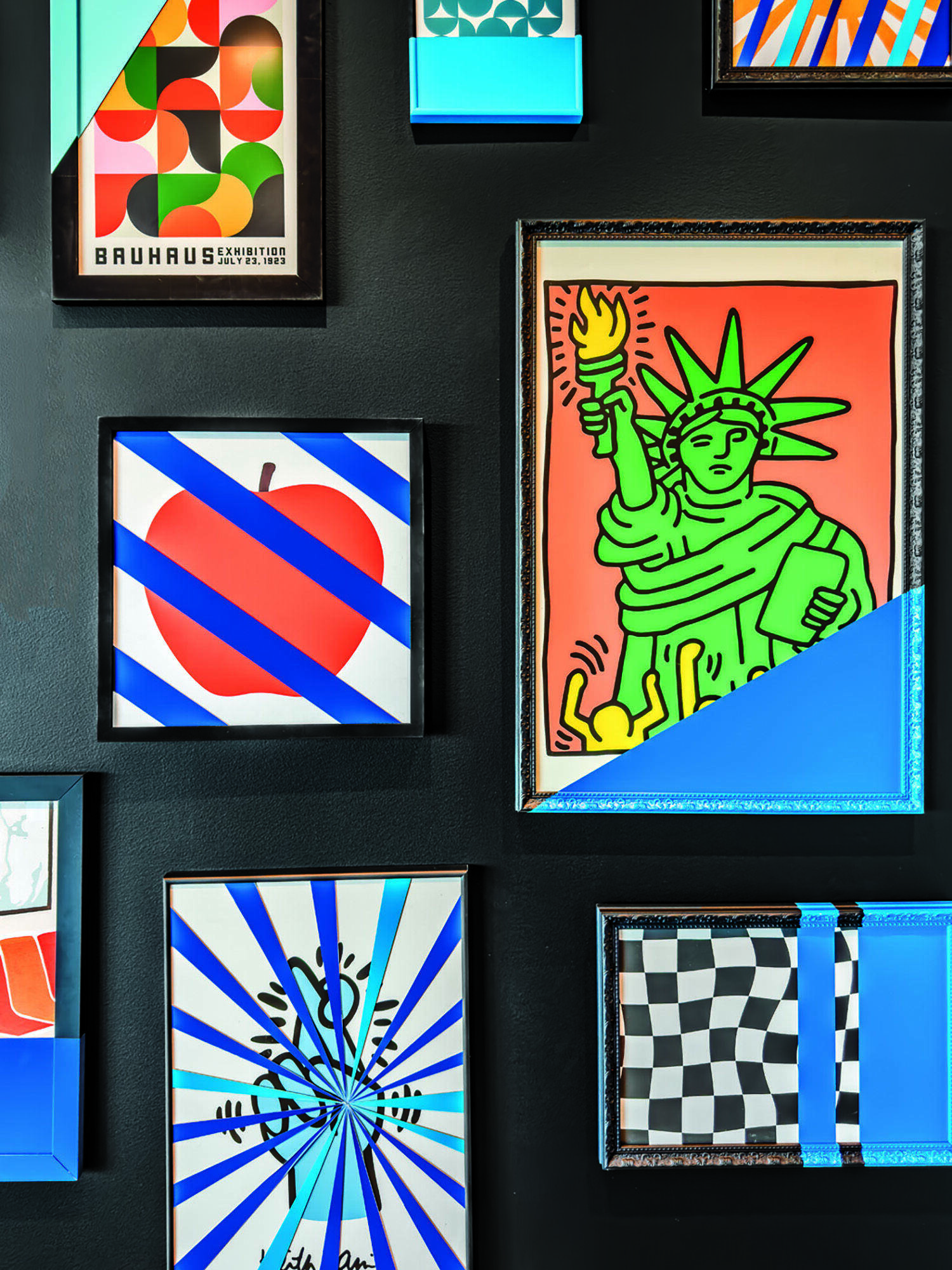 A black wall displays colorful framed artworks, including abstract patterns, an apple, the Statue of Liberty, and blue tape accents across several frames.