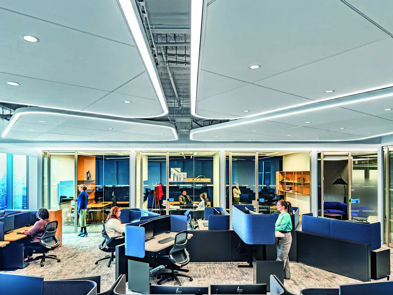 Modern office with several people working at desks and standing, open cubicles, large windows, and bright ceiling lights.