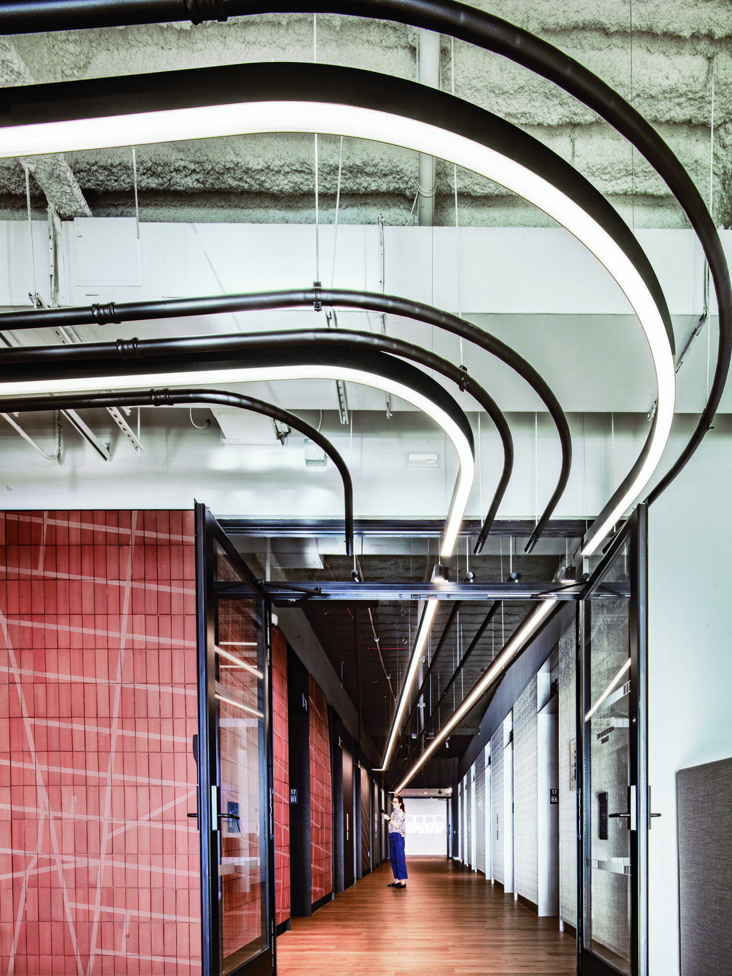 Modern office hallway with curved overhead LED lights, glass office doors on both sides, red tile walls on the left, and a person standing in the distance.