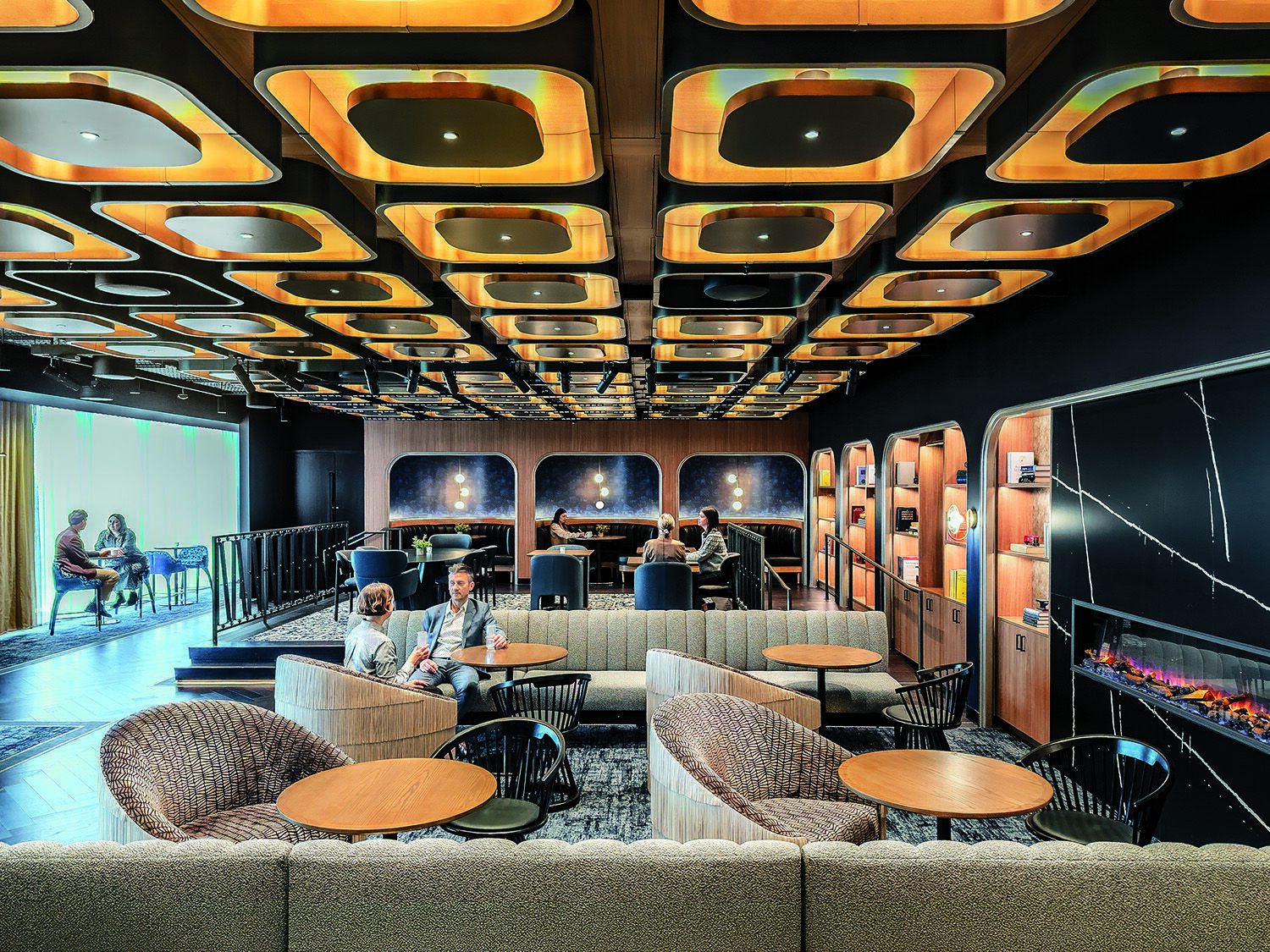 Modern lounge area with stylish wooden ceiling panels, cozy seating, round tables, built-in shelves, and an electric fireplace. Several people are seated and conversing throughout the space.