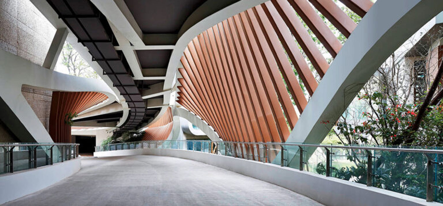 Curved concrete walkway with a glass railing, sheltered by a rhythmic structure of angled vertical wooden slats.
