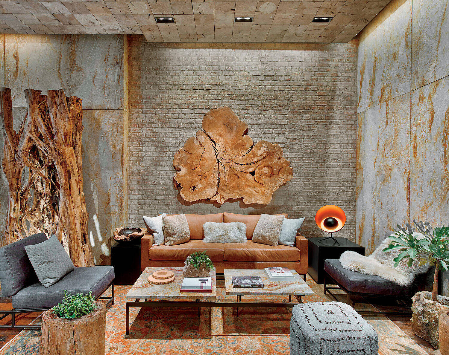 Modern living room with a tan leather sofa, wooden wall art, stone walls, two gray chairs, plants, and a round orange lamp on a side table.