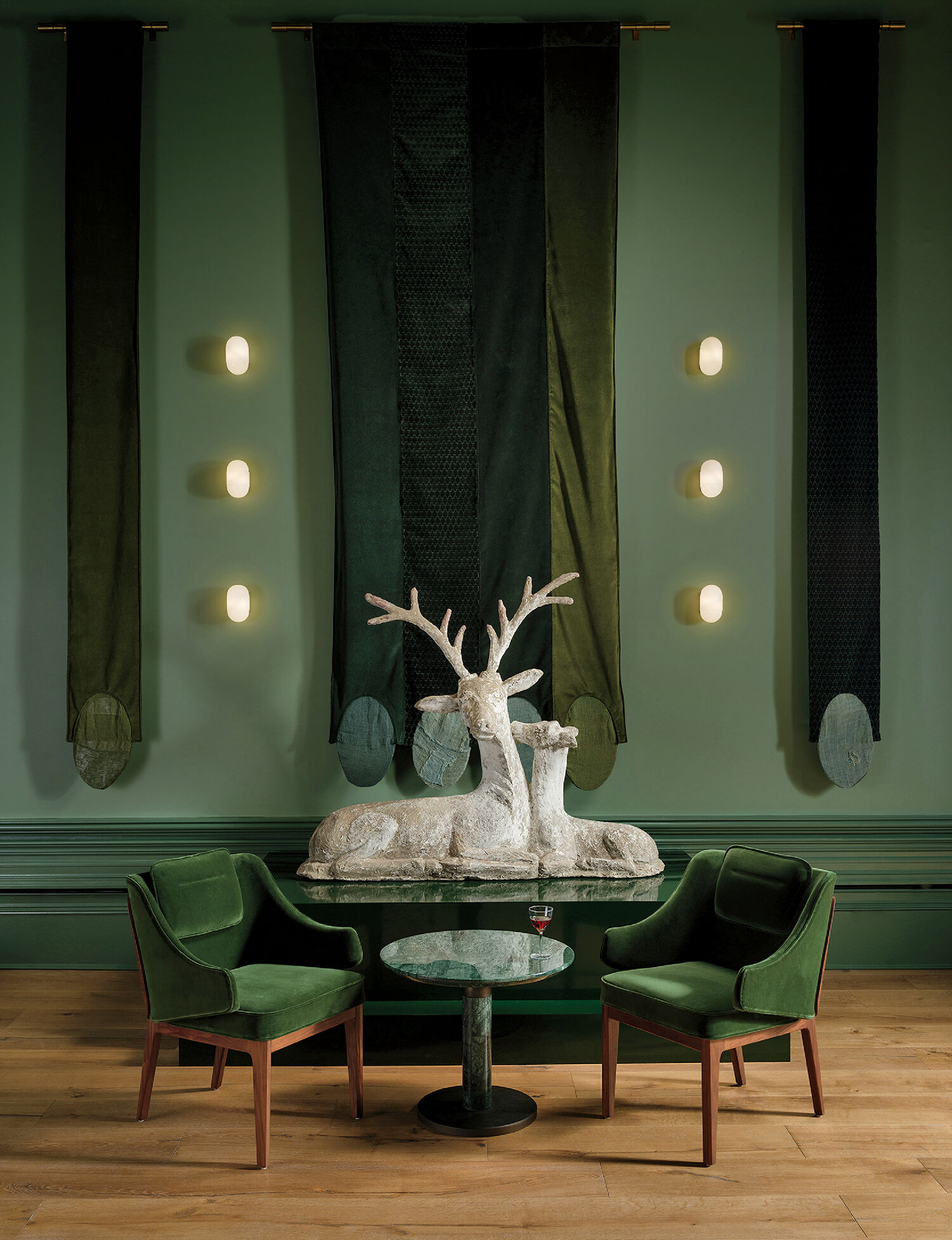 Sculptures of two deer on a small round table, flanked by two green velvet chairs, with dark green banners and wall lights on a green wall.