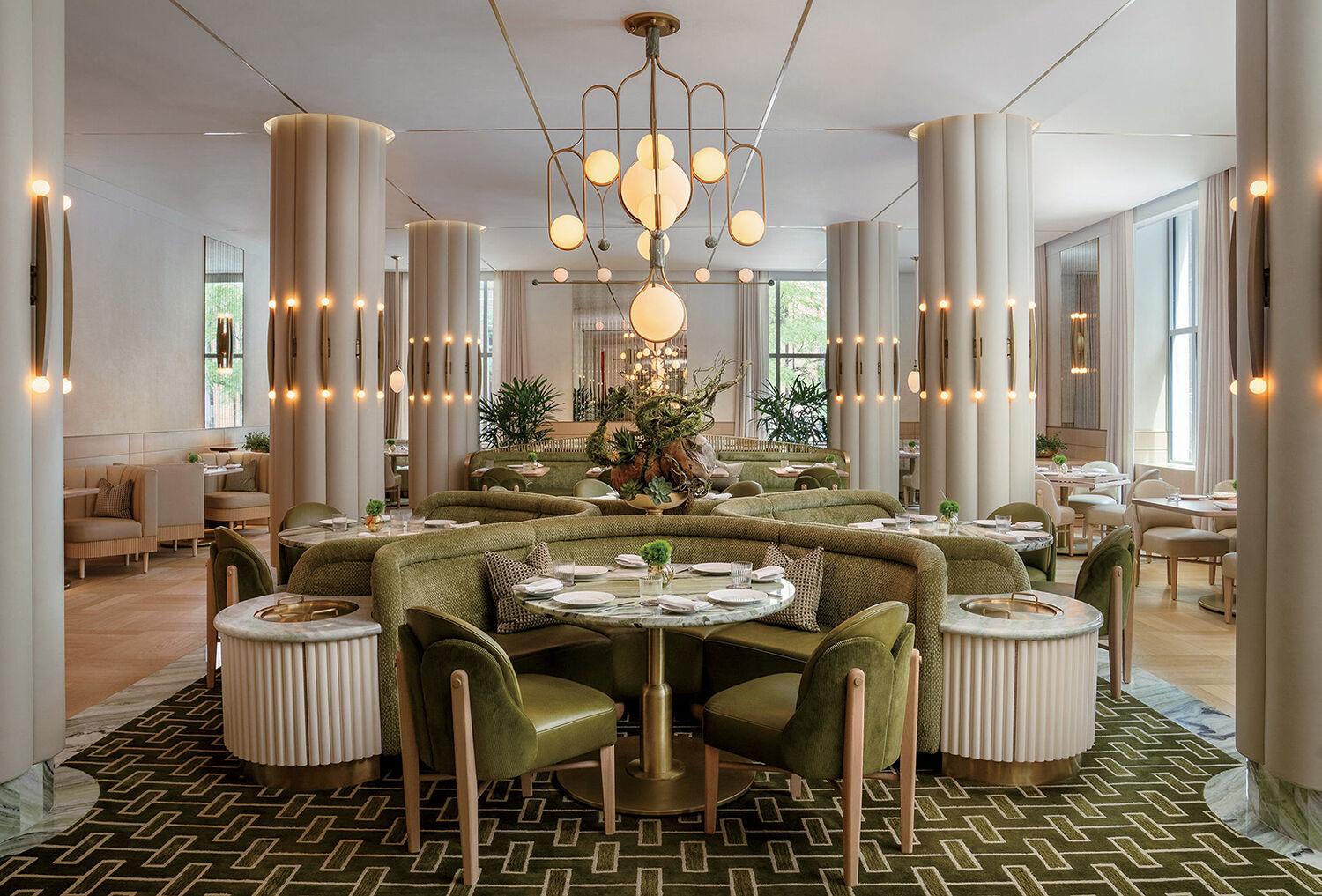 Elegant restaurant interior with green velvet booths, round tables set for dining, modern pendant lights, and decorative columns. Large windows and indoor plants add brightness to the space.