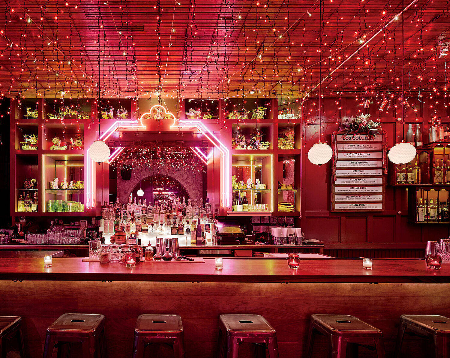 A bar with a wooden counter, metal stools, illuminated bottles, red string lights on the ceiling, and a neon-lit shelving unit behind the bar.