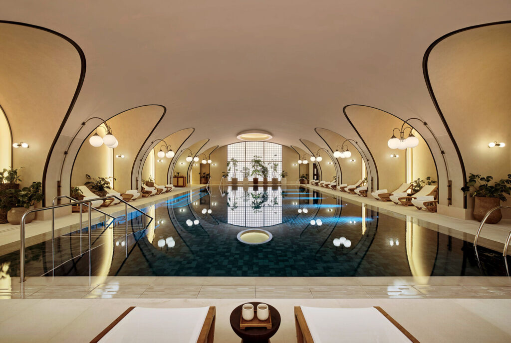 Indoor swimming pool with modern, arched ceiling, lounge chairs along the sides, potted plants, and soft lighting creating a calm, symmetrical atmosphere.