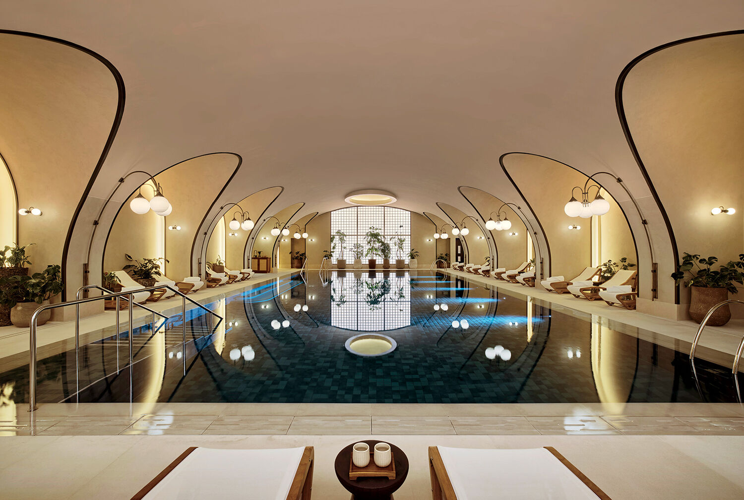 Indoor swimming pool with modern, arched ceiling, lounge chairs along the sides, potted plants, and soft lighting creating a calm, symmetrical atmosphere.