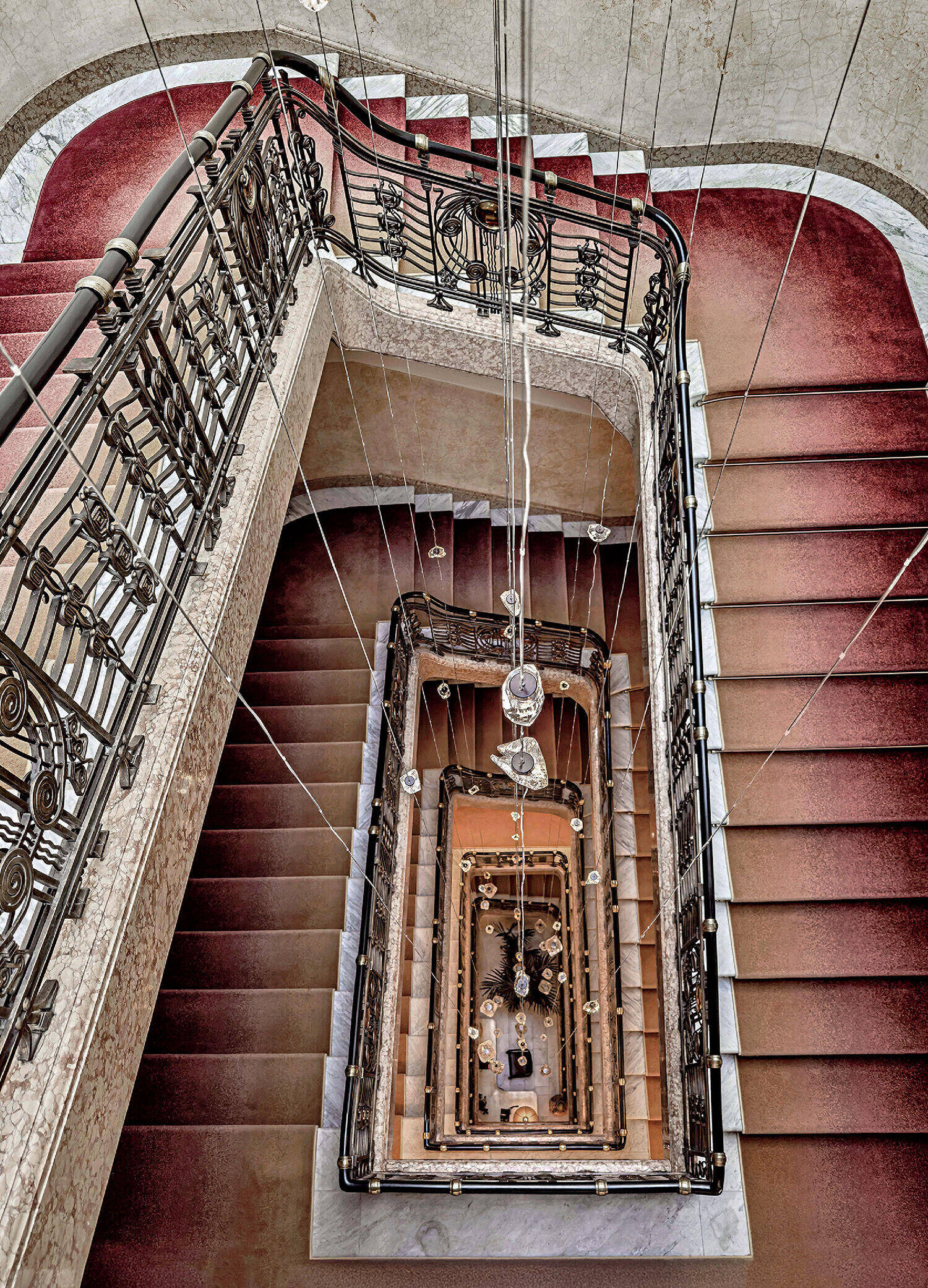 A spiral staircase with ornate railings and red carpet, viewed from above, with hanging pendant lights descending through the stairwell.