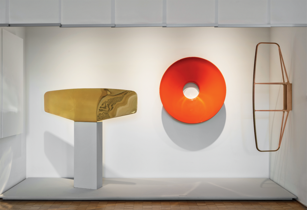 Three abstract sculptures are displayed in a minimalist white space: a gold rectangular form, a red circular disc with a hole, and a copper wireframe structure.