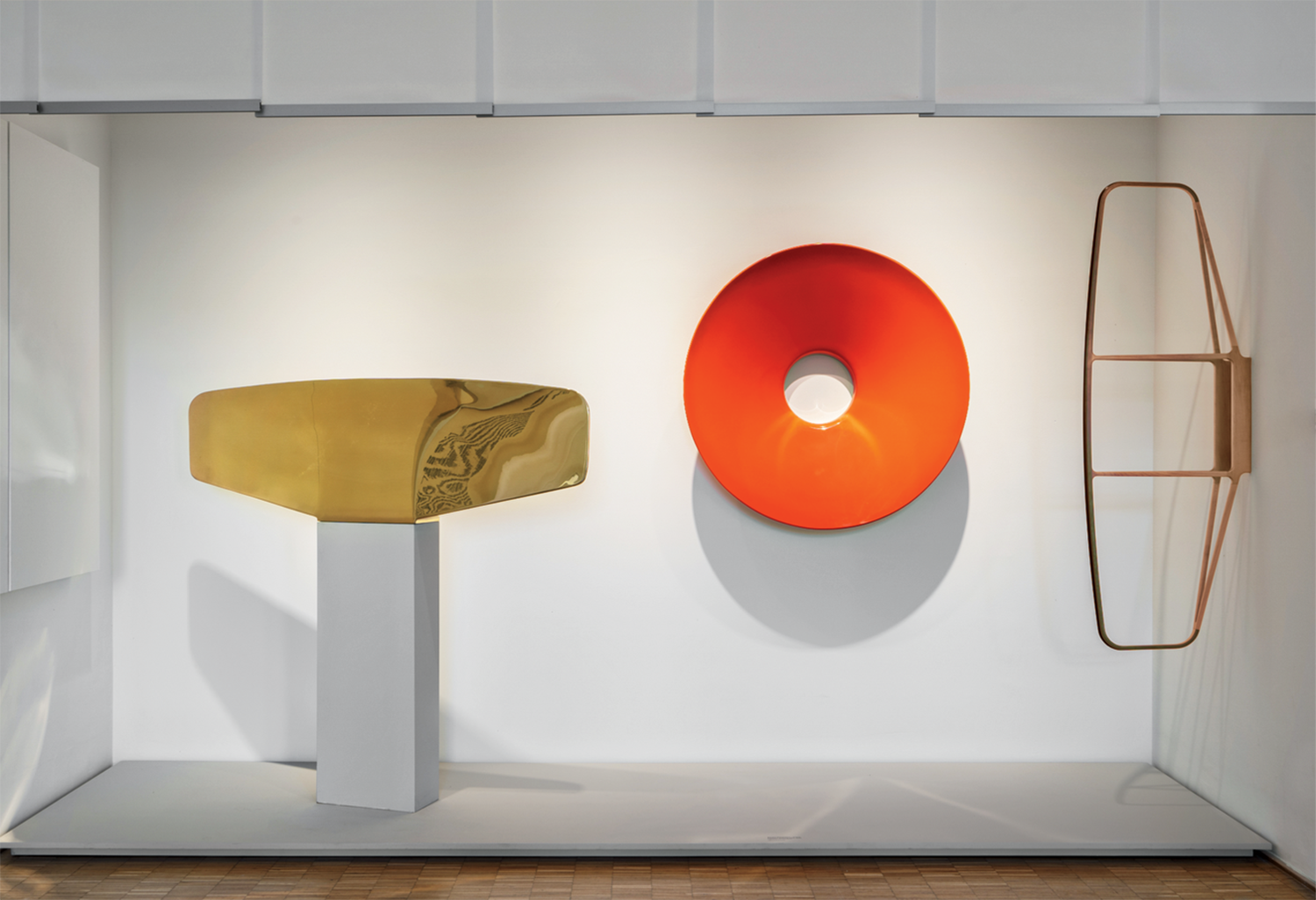 Three abstract sculptures are displayed in a minimalist white space: a gold rectangular form, a red circular disc with a hole, and a copper wireframe structure.